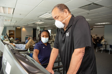 Masked female therapist supervising masked male patient walking on treadmill