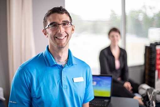 male therapist wearing a teal blue shirt and name tag standing in front of a laptop on a desk